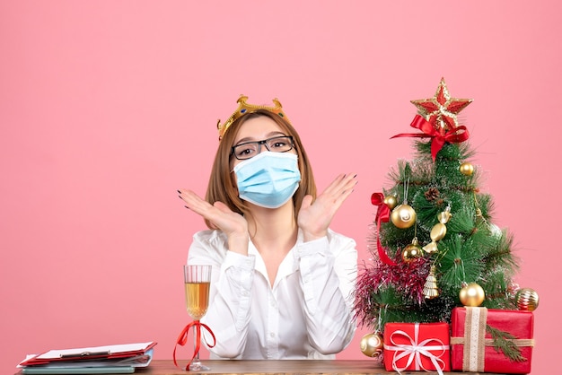 Foto gratuita vista frontal de la trabajadora en máscara sentada detrás de su mesa con regalos de navidad en rosa