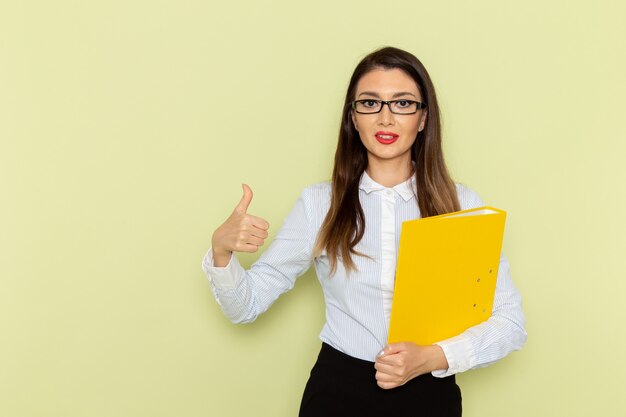 Vista frontal de la trabajadora de oficina en camisa blanca y falda negra sosteniendo un archivo amarillo y sonriendo en la pared verde