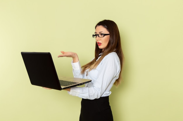 Vista frontal de la trabajadora de oficina en camisa blanca y falda negra sosteniendo su computadora portátil en la pared verde