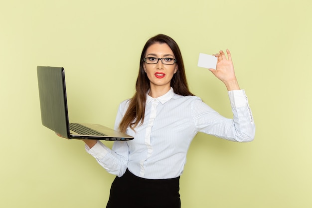 Vista frontal de la trabajadora de oficina con camisa blanca y falda negra usando su computadora portátil y sosteniendo una tarjeta en la pared verde claro