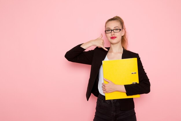 Vista frontal de la trabajadora de oficina en chaqueta negra estricta con archivo amarillo sonriendo en la pared rosa claro