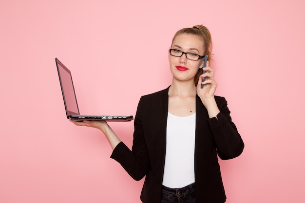 Vista frontal de la trabajadora de oficina en chaqueta negra estricta hablando por teléfono y usando la computadora portátil en la pared rosa