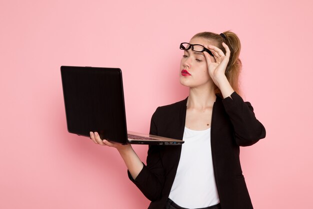 Vista frontal de la trabajadora de oficina en chaqueta negra estricta con laptop en pared rosa claro