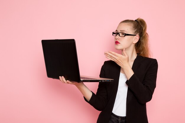 Vista frontal de la trabajadora de oficina en chaqueta negra estricta sosteniendo su computadora portátil en la pared rosa claro