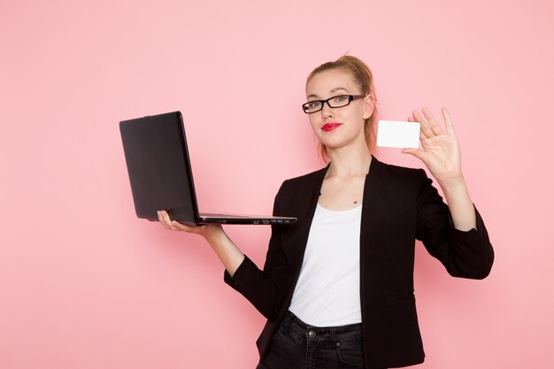 Vista frontal de la trabajadora de oficina en chaqueta negra estricta sosteniendo la tarjeta y usando su computadora portátil en la pared rosa