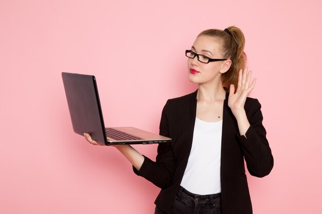 Vista frontal de la trabajadora de oficina en chaqueta negra estricta sosteniendo y usando su computadora portátil en la pared rosa