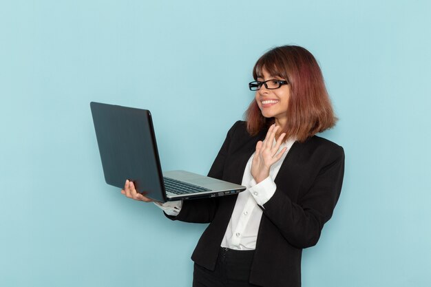 Vista frontal de la trabajadora de oficina en traje estricto sosteniendo el portátil usándolo en la superficie azul