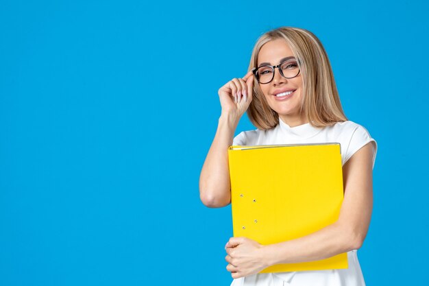 Vista frontal de la trabajadora en vestido blanco sosteniendo una carpeta amarilla en la pared azul