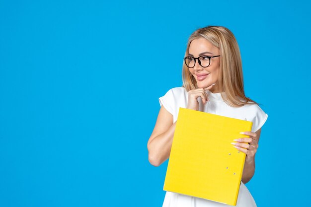 Vista frontal de la trabajadora en vestido blanco sosteniendo una carpeta amarilla y sonriendo en la pared azul