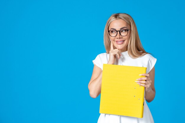 Vista frontal de la trabajadora en vestido blanco sosteniendo una carpeta amarilla y sonriendo en la pared azul