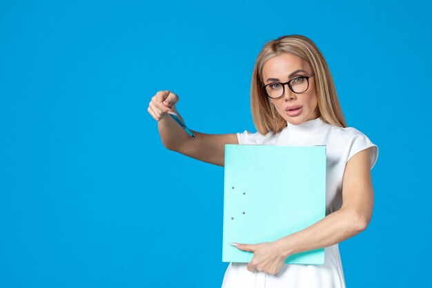 Vista frontal de la trabajadora en vestido blanco sosteniendo la carpeta en la pared azul