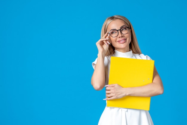 Vista frontal de la trabajadora en vestido blanco sosteniendo una carpeta con una sonrisa en la pared azul