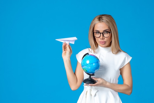 Vista frontal de la trabajadora en vestido blanco sosteniendo globo terráqueo y avión de papel en la pared azul