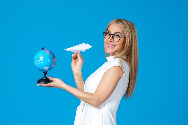 Vista frontal de la trabajadora en vestido blanco sosteniendo globo terráqueo y avión de papel en la pared azul