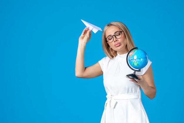 Vista frontal de la trabajadora en vestido blanco sosteniendo globo terráqueo y avión de papel en la pared azul