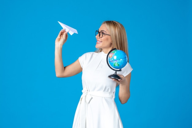 Vista frontal de la trabajadora en vestido blanco sosteniendo globo terráqueo y avión de papel en la pared azul