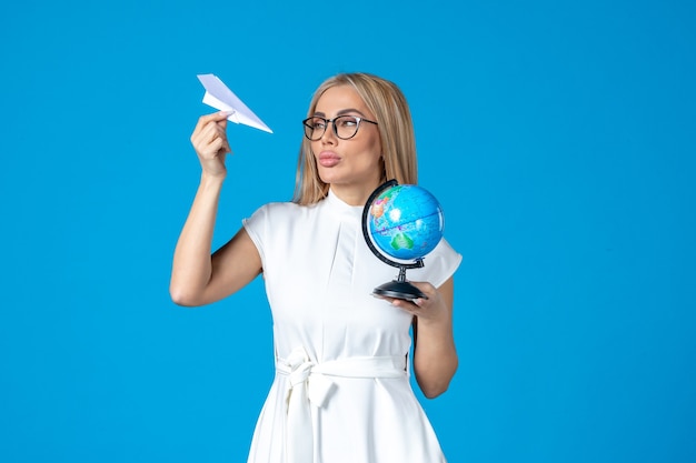 Foto gratuita vista frontal de la trabajadora en vestido blanco sosteniendo globo terráqueo y avión de papel en la pared azul