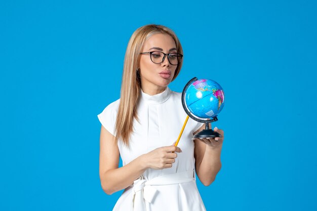 Vista frontal de la trabajadora en vestido blanco sosteniendo un pequeño globo terráqueo en la pared azul