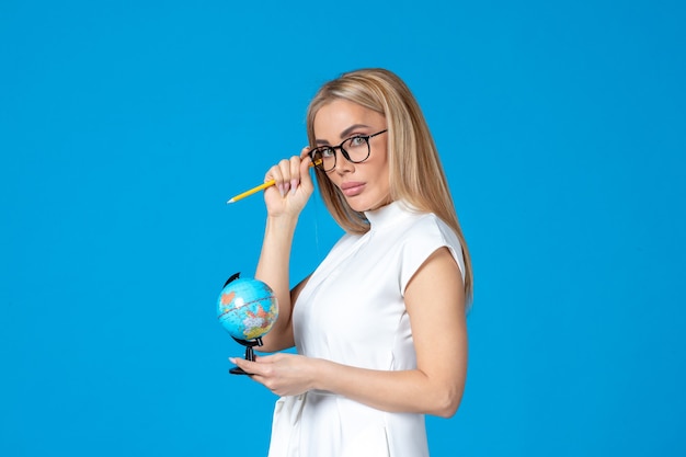 Vista frontal de la trabajadora en vestido blanco sosteniendo un pequeño globo terráqueo en la pared azul