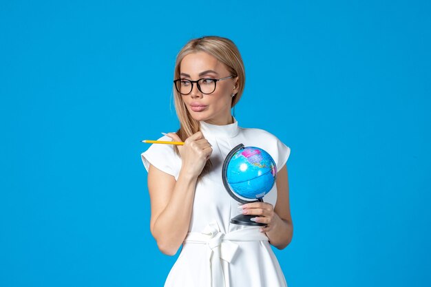 Vista frontal de la trabajadora en vestido blanco sosteniendo un pequeño globo terráqueo en la pared azul