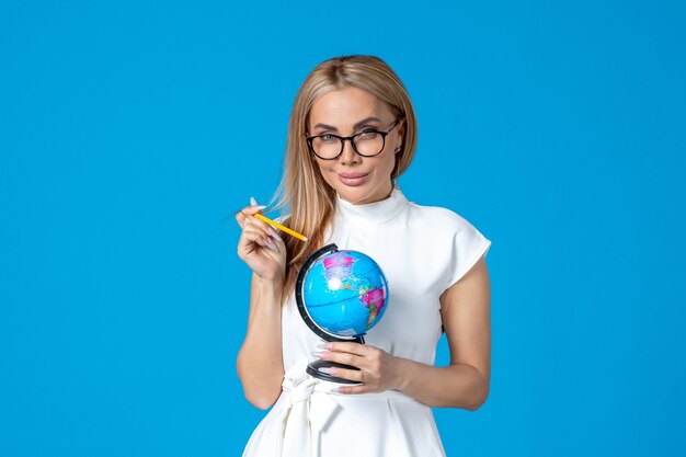 Vista frontal de la trabajadora en vestido blanco sosteniendo un pequeño globo terráqueo en la pared azul