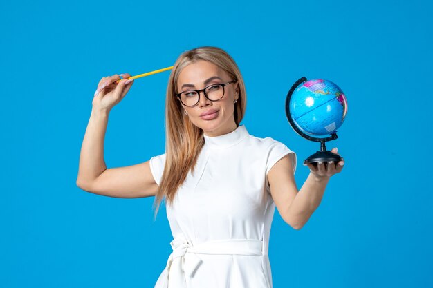 Vista frontal de la trabajadora en vestido blanco sosteniendo un pequeño globo terráqueo en la pared azul