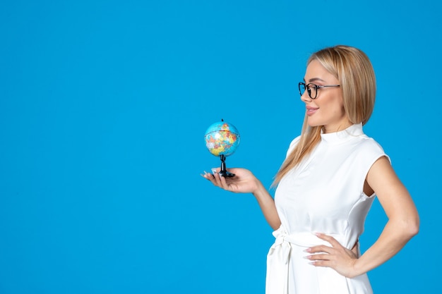 Vista frontal de la trabajadora en vestido blanco sosteniendo un pequeño globo terráqueo en la pared azul