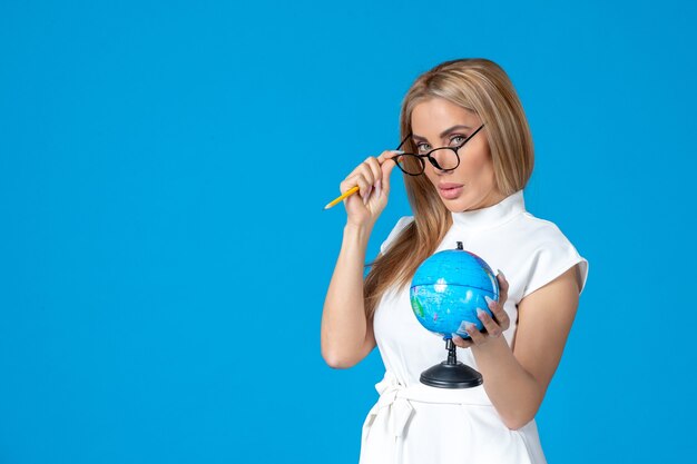 Vista frontal de la trabajadora en vestido blanco sosteniendo un pequeño globo terráqueo en la pared azul