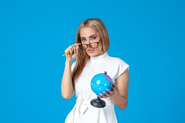 Vista frontal de la trabajadora en vestido blanco sosteniendo un pequeño globo terráqueo en la pared azul