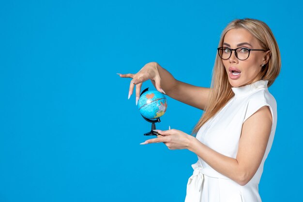 Vista frontal de la trabajadora en vestido blanco sosteniendo un pequeño globo terráqueo en la pared azul