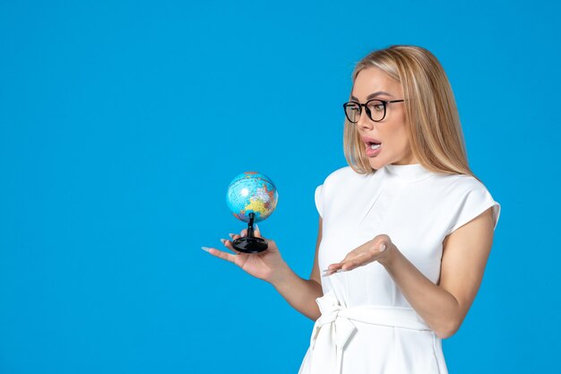 Vista frontal de la trabajadora en vestido blanco sosteniendo un pequeño globo terráqueo en la pared azul