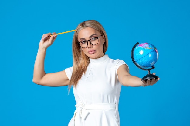 Vista frontal de la trabajadora en vestido blanco sosteniendo un pequeño globo terráqueo en la pared azul