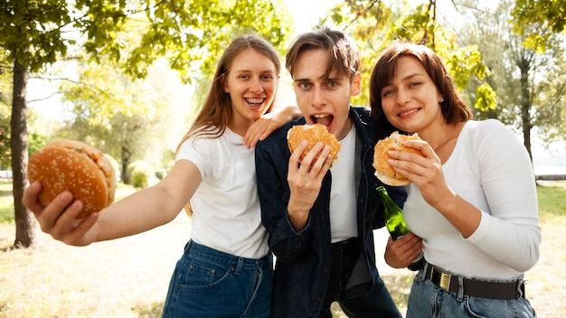 Vista frontal de tres amigos en el parque con cerveza y hamburguesas.