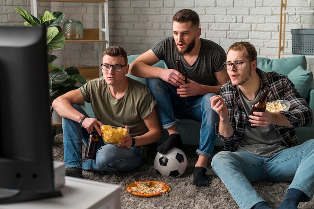 Vista frontal de tres amigos varones viendo deportes en la televisión juntos mientras toman bocadillos y cerveza