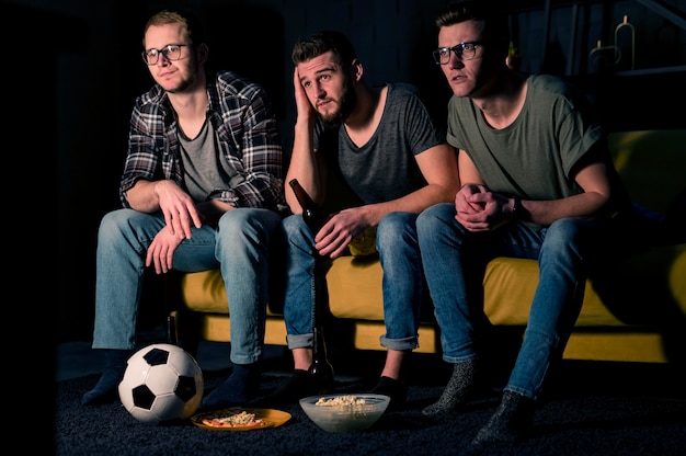 Foto gratuita vista frontal de tres amigos varones viendo deportes en la televisión juntos mientras toman bocadillos y cerveza