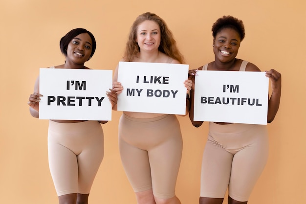 Vista frontal de tres mujeres sosteniendo pancartas con declaraciones de positividad corporal