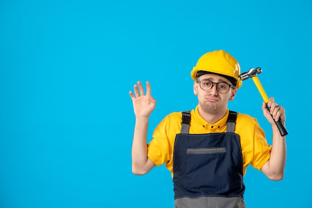 Foto gratuita vista frontal del triste constructor masculino en uniforme con un martillo en sus manos sobre un azul