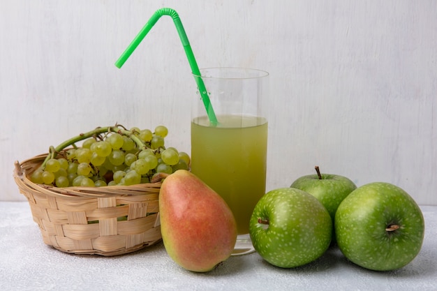 Vista frontal de uvas verdes en una canasta con pera manzanas verdes y jugo de manzana con una pajita verde en un vaso sobre un fondo blanco.