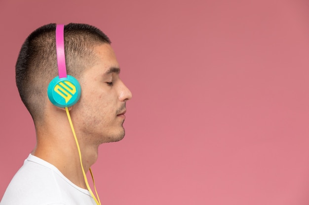 Vista frontal del varón joven en camiseta blanca escuchando música a través de auriculares en el fondo rosa claro