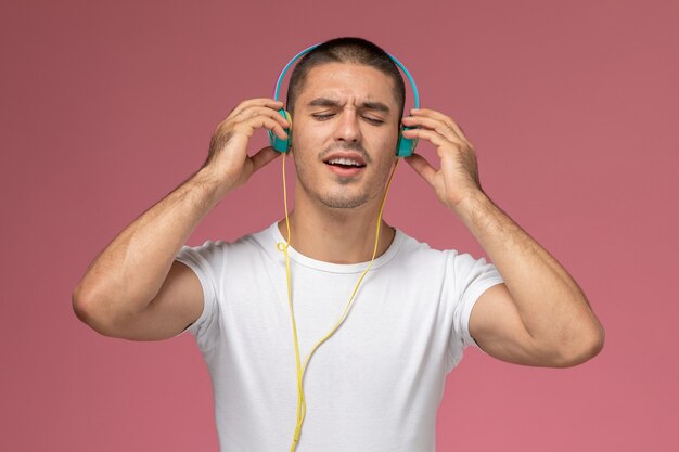 Vista frontal del varón joven en camiseta blanca escuchando música a través de auriculares en el fondo rosa