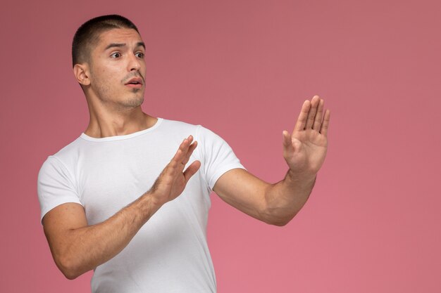 Vista frontal del varón joven en camiseta blanca posando con expresión cautelosa sobre fondo rosa