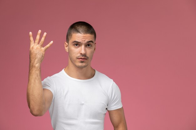 Vista frontal del varón joven en camiseta blanca posando mostrando cuatro signos sobre fondo rosa