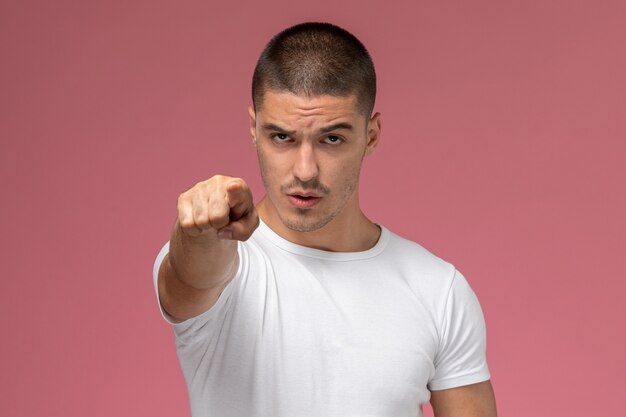 Vista frontal del varón joven en camiseta blanca posando señalando sobre fondo rosa