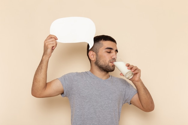 Vista frontal del varón joven en camiseta gris bebiendo leche y sosteniendo un cartel blanco sobre beige