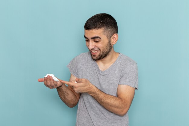 Vista frontal del varón joven en camiseta gris con espuma blanca para afeitarse en la barba azul hielo espuma afeitadora de pelo