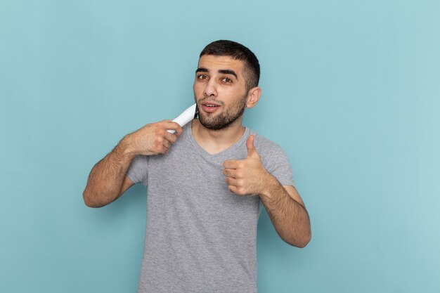 Vista frontal del varón joven en camiseta gris con maquinilla de afeitar eléctrica en azul hielo barba de afeitar espuma de pelo masculino