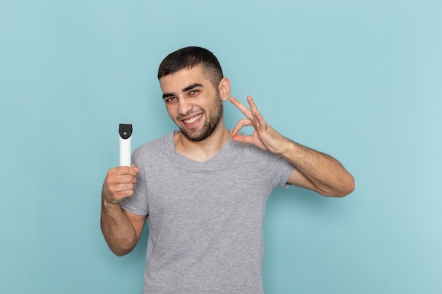 Vista frontal del varón joven en camiseta gris con maquinilla de afeitar eléctrica con sonrisa en el azul