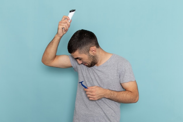 Vista frontal del varón joven en camiseta gris con maquinilla de afeitar y maquinilla de afeitar eléctrica en azul claro