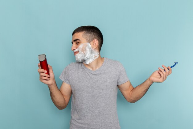 Vista frontal del varón joven en camiseta gris sosteniendo una maquinilla de afeitar eléctrica y simple en la barba azul hielo macho de afeitar con espuma