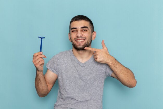 Vista frontal del varón joven en camiseta gris sosteniendo la navaja y sonriendo en la barba de afeitar azul